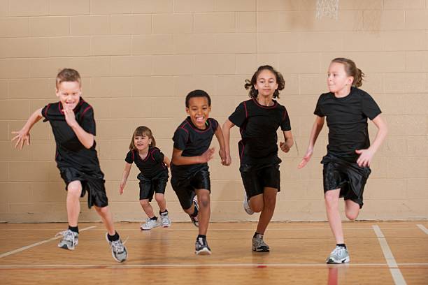 Children running on a gym floor.  The children are all wearing black shirts and black shorts.  Their tennis shoes are different colors and their socks are black.  The gym floor is light brown with white lines in various places.  The children are different sizes and ages.